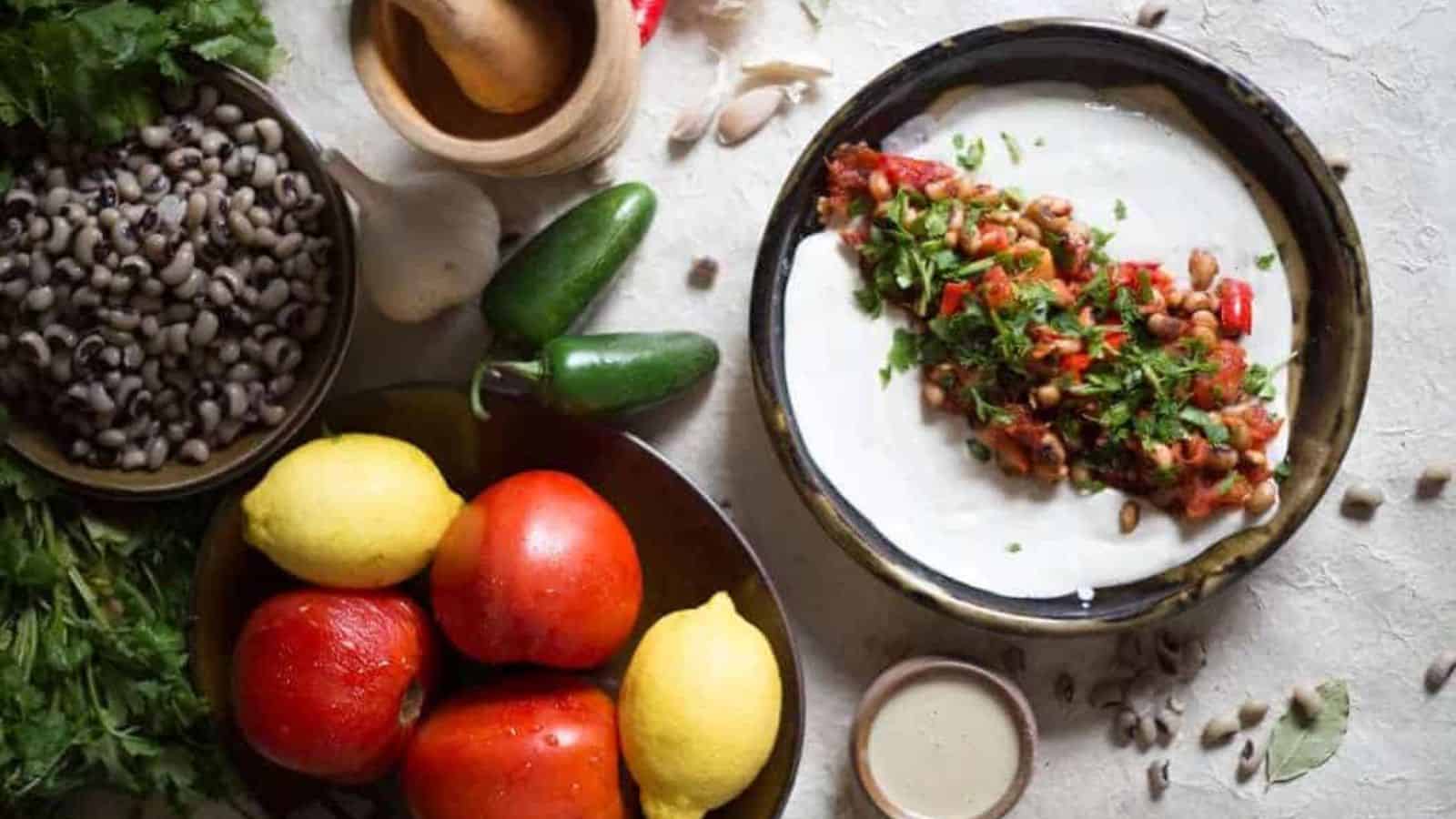 A bowl with a creamy white spread topped with a bean and tomato mixture, surrounded by fresh lemons, tomatoes, jalapeños, herbs, a wooden mortar and pestle, garlic, and a small bowl of sauce.