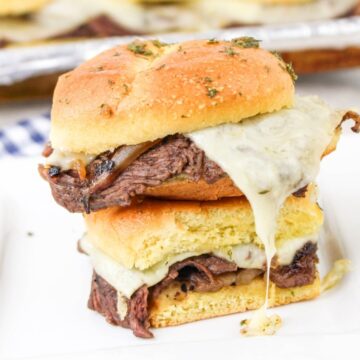 A sandwich with sliced beef, melted cheese, and grilled onions is stacked on a white plate, with another sandwich partially visible on top. The sandwiches sit on a patterned cloth with a tray in the background.