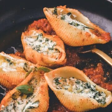 Five stuffed pasta shells filled with ricotta and spinach rest on a bed of tomato sauce in a black dish. A fork is placed on the plate. The dish is garnished with herbs.