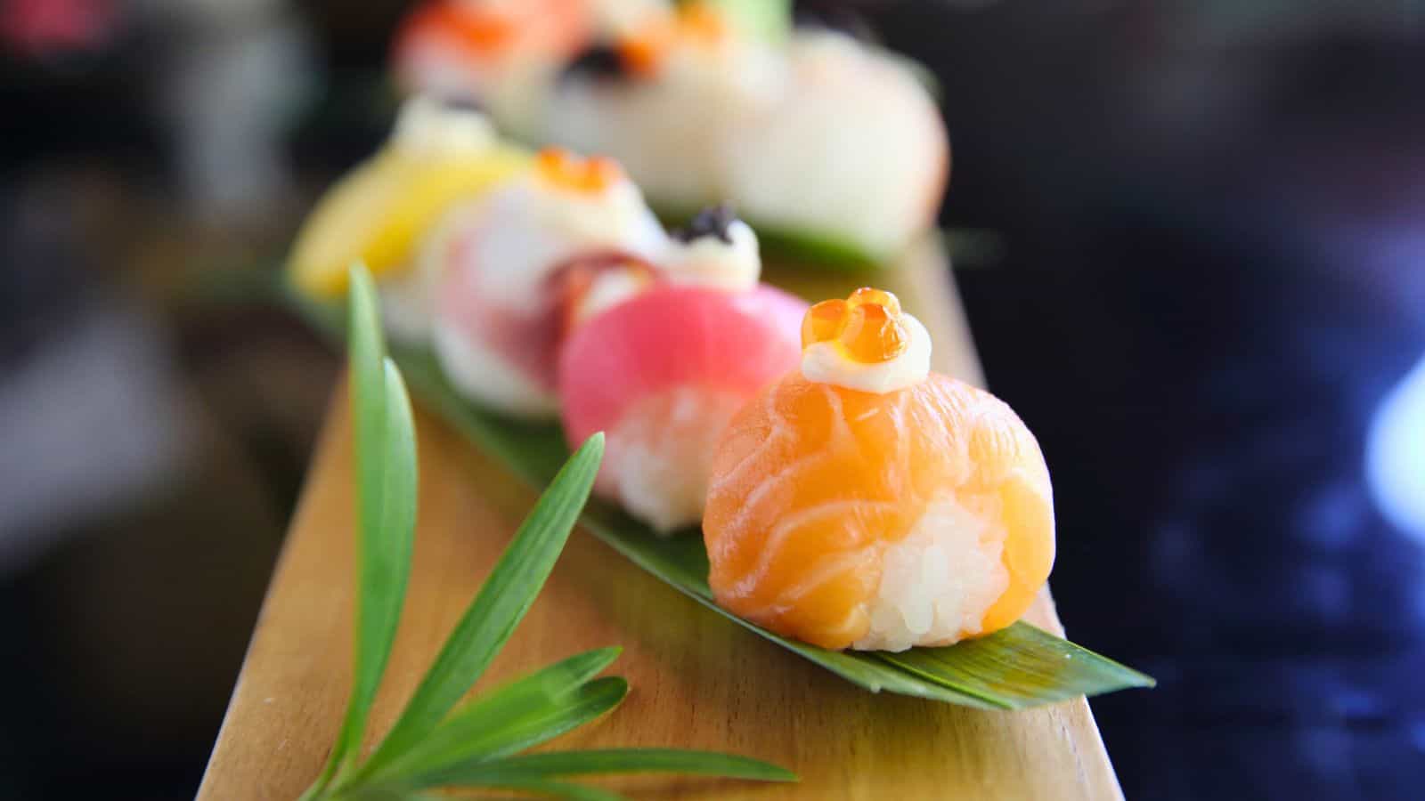 A row of sushi bites sits on a wooden board, each topped with different fish and garnishes. The front piece has a slice of salmon with a small dollop of cream and an orange roe on top. A green leaf decorates the board.