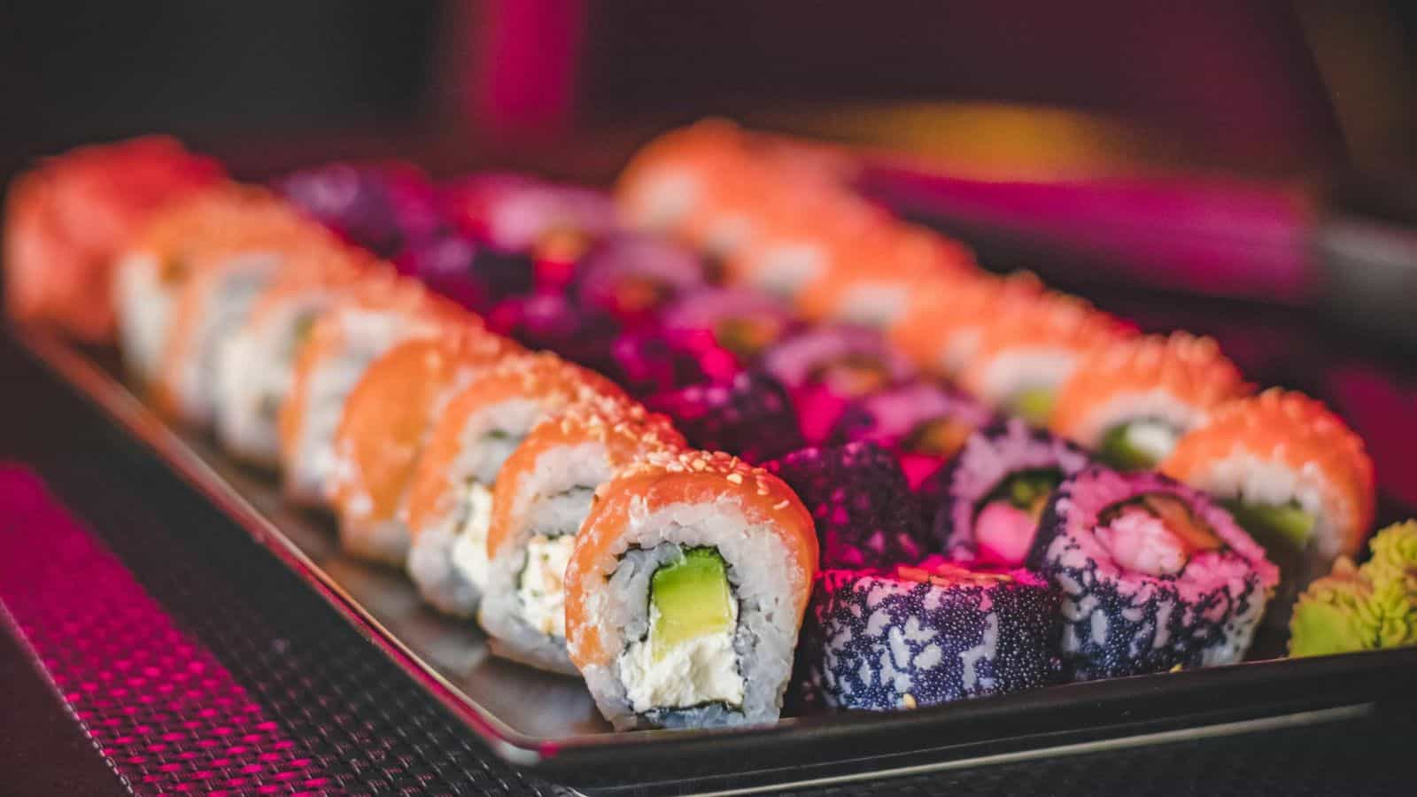 A tray of assorted sushi rolls is displayed. Some rolls are topped with orange fish and sesame seeds, while others feature dark and colorful patterns. Cream cheese and avocado are visible in some pieces. Wasabi is placed on the side.