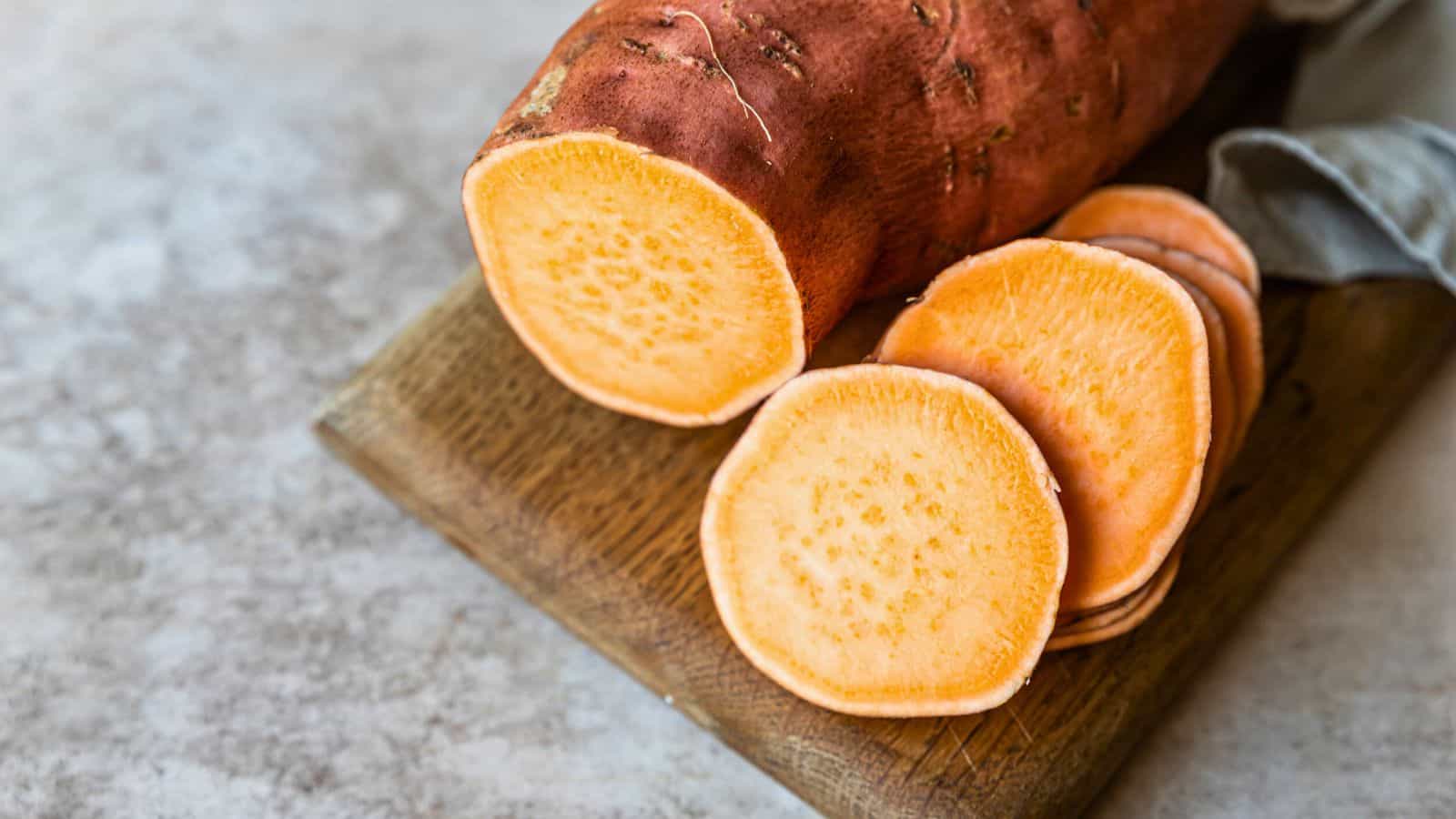 A whole sweet potato and several round slices are arranged on a wooden cutting board. The sweet potato has an orange interior and a reddish-brown skin. A piece of blue-gray cloth is partially visible to the right.