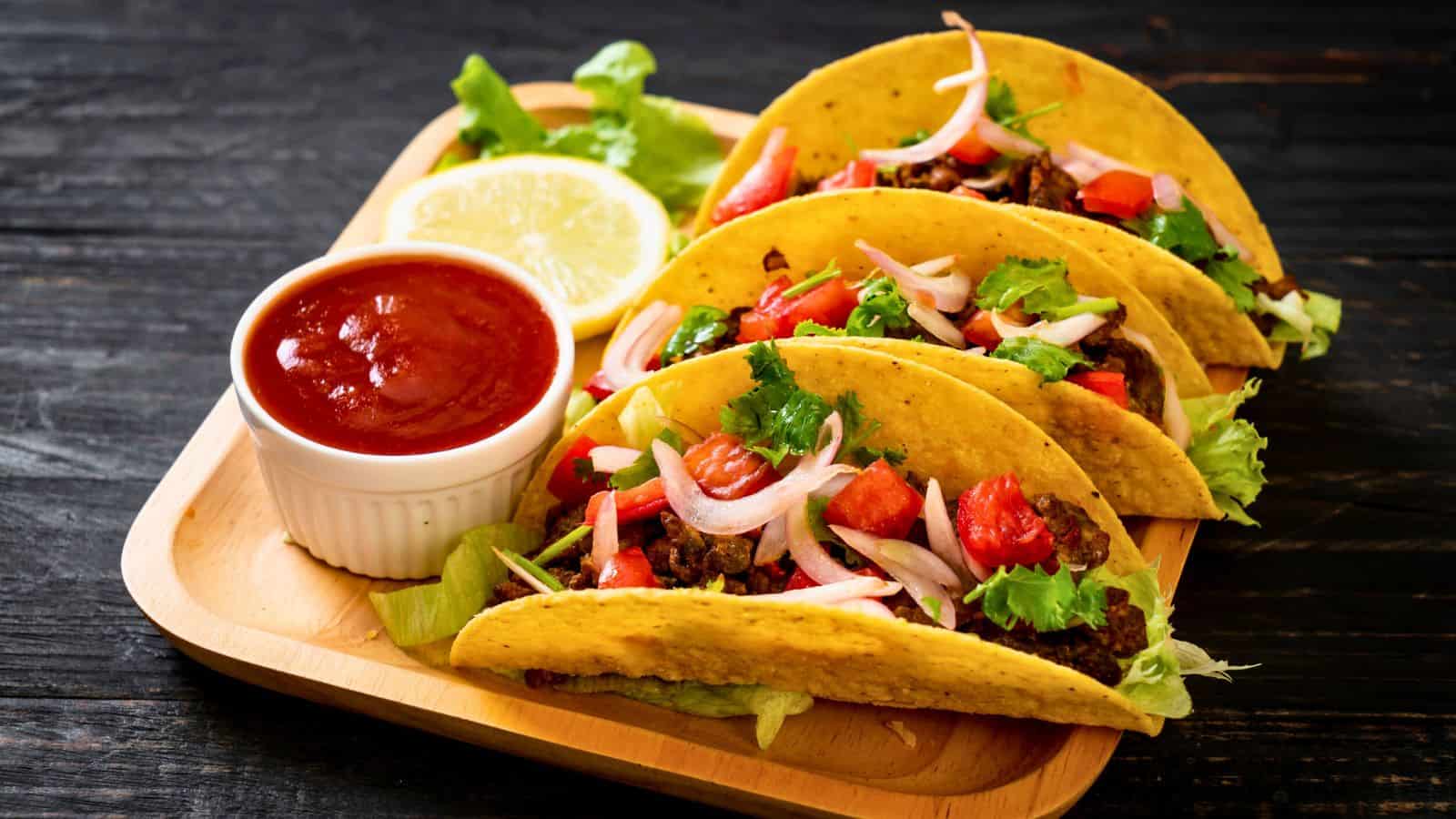 A wooden tray holds three tacos with yellow shells, filled with lettuce, diced tomatoes, sliced onions, and cilantro. A small cup of red sauce and a lemon wedge are placed alongside the tacos. The background is a dark surface.