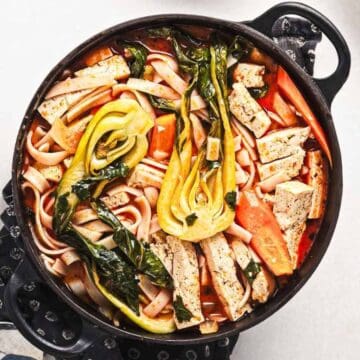 A top-down view of a pot filled with noodle soup containing bok choy, sliced tofu, carrots, and leafy greens. The pot is placed on a black pot holder on a light-colored surface.