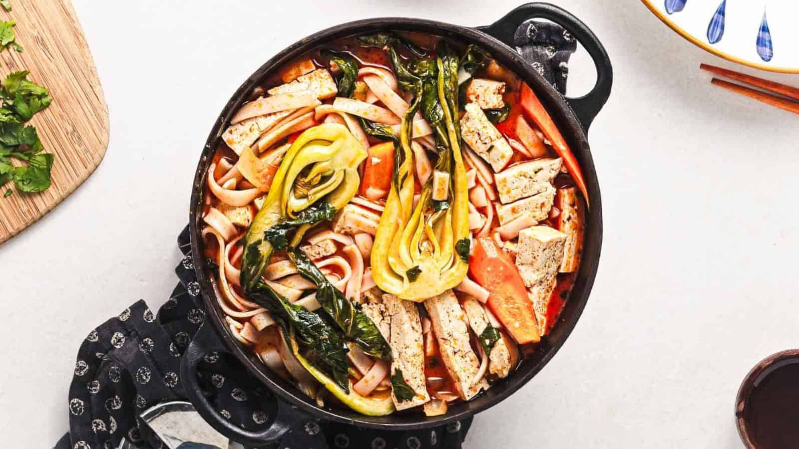 A bowl of noodle soup with tofu strips, baby bok choy, carrot slices, and fresh herbs, set on a white surface with a spoon nearby and a small bowl of chopped greens in the background.