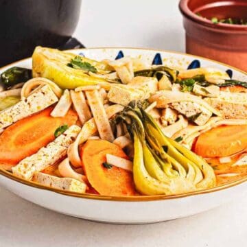 A bowl of noodle soup with tofu strips, baby bok choy, carrot slices, and fresh herbs, set on a white surface with a spoon nearby and a small bowl of chopped greens in the background.