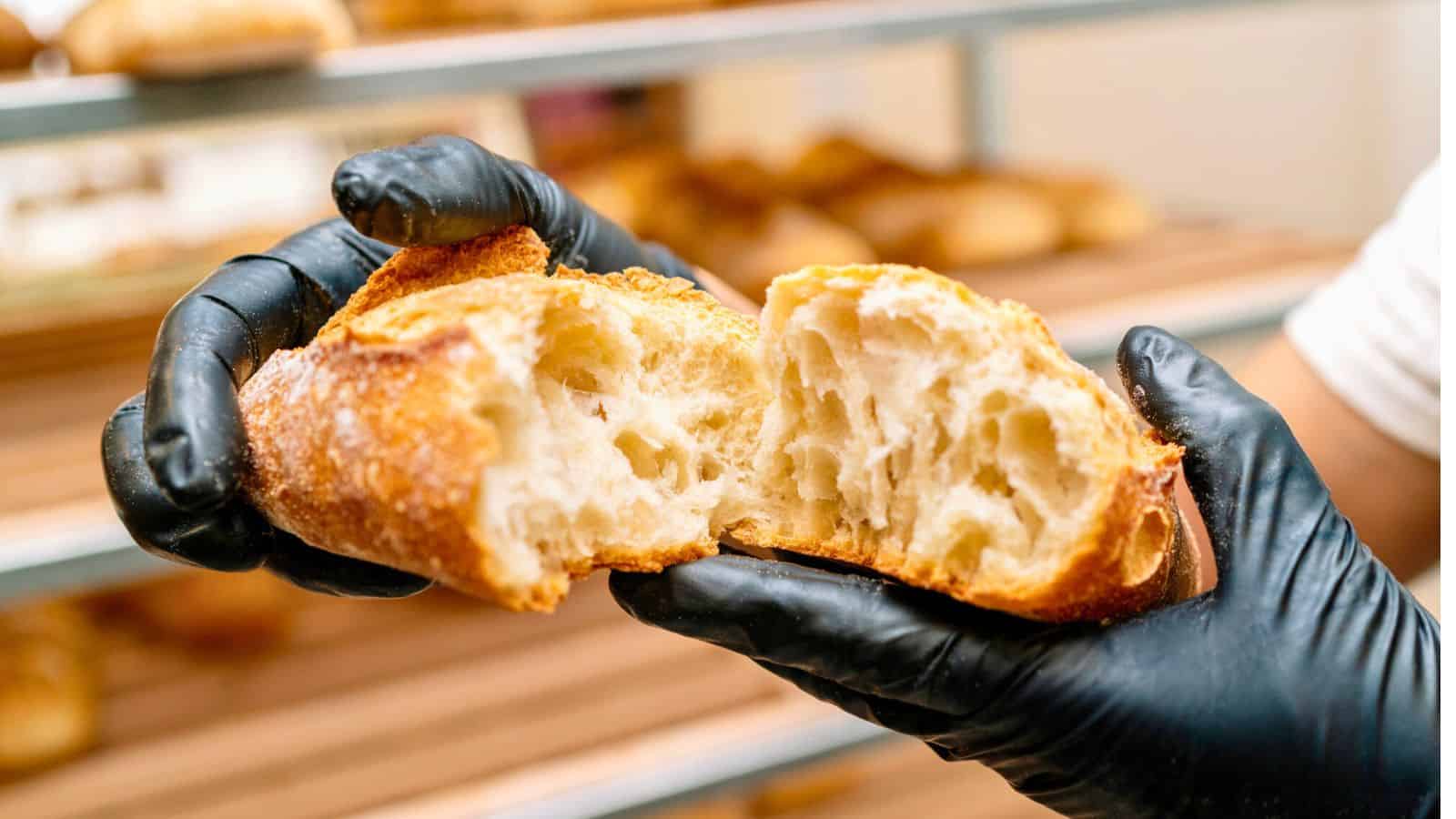 A person wearing black gloves holds a freshly baked baguette broken in half, revealing its airy interior. The background shows shelves with more bread.