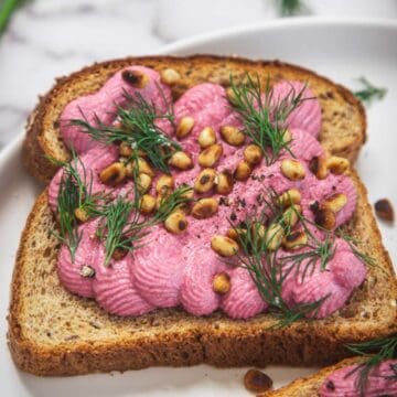 A slice of whole grain toast topped with creamy beet spread, toasted pine nuts, and fresh dill, served on a white plate.