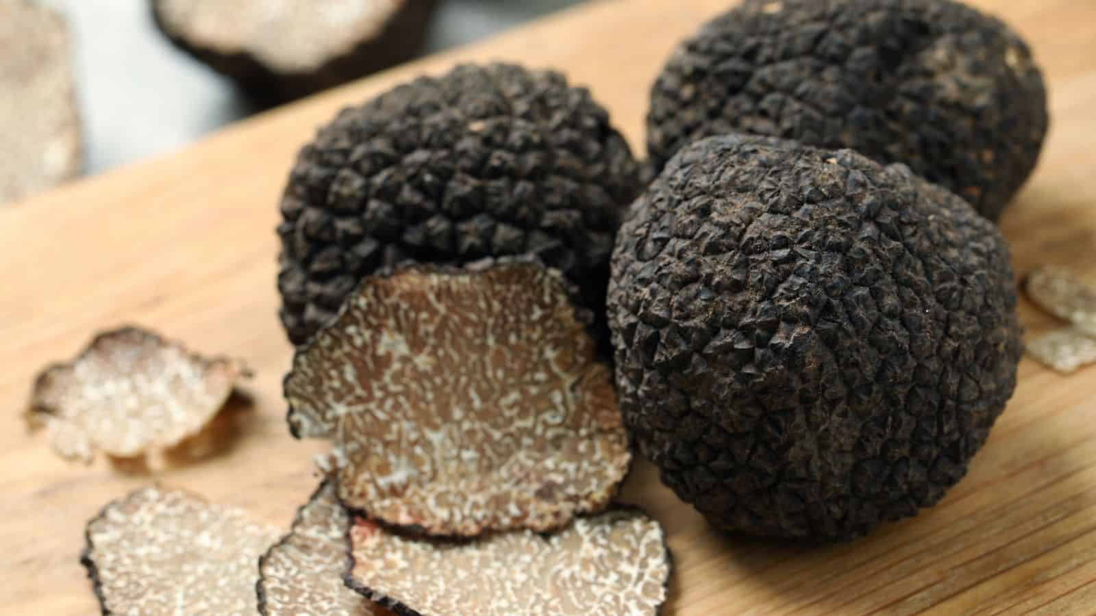 Whole black truffles and several slices are displayed on a wooden surface. The truffles have a rough, textured exterior, while the sliced pieces reveal a marbled interior pattern. The setting appears to be a kitchen or food preparation area.