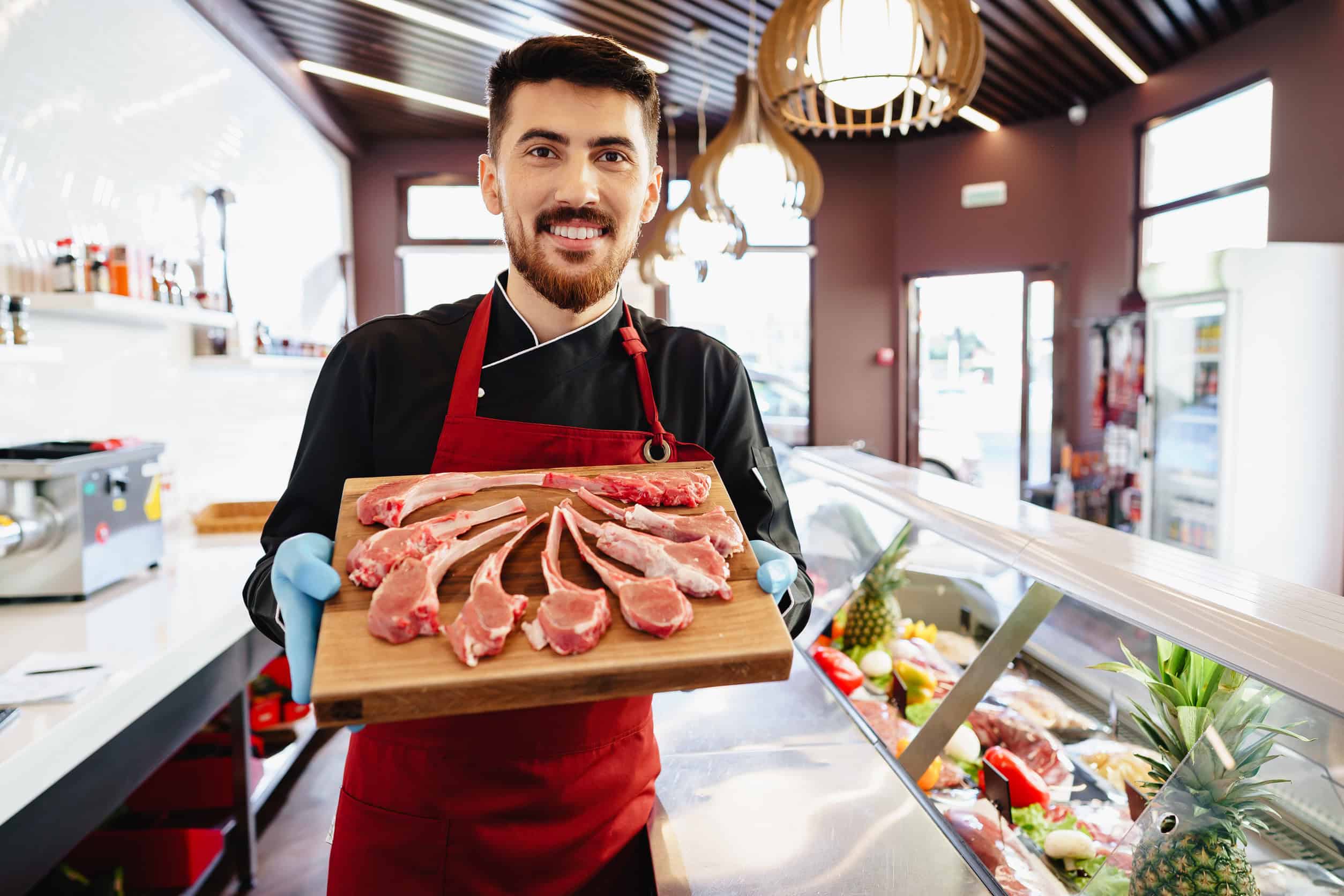 A butcher in a red apron and black shirt holds a wooden board with raw lamb chops, showcasing quality meat for those seeking 15 smart ways to save money on food. He smiles confidently in his modern shop, where various meats and products are displayed enticingly in the background.