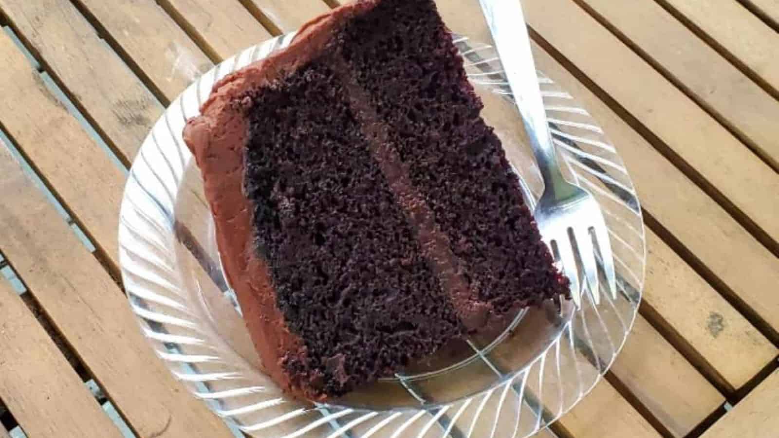 A slice of chocolate layer cake with chocolate frosting sits on a clear glass plate beside a fork, atop a wooden slatted surface—perfect for Mother’s Day desserts or as one of the special cakes for mom.