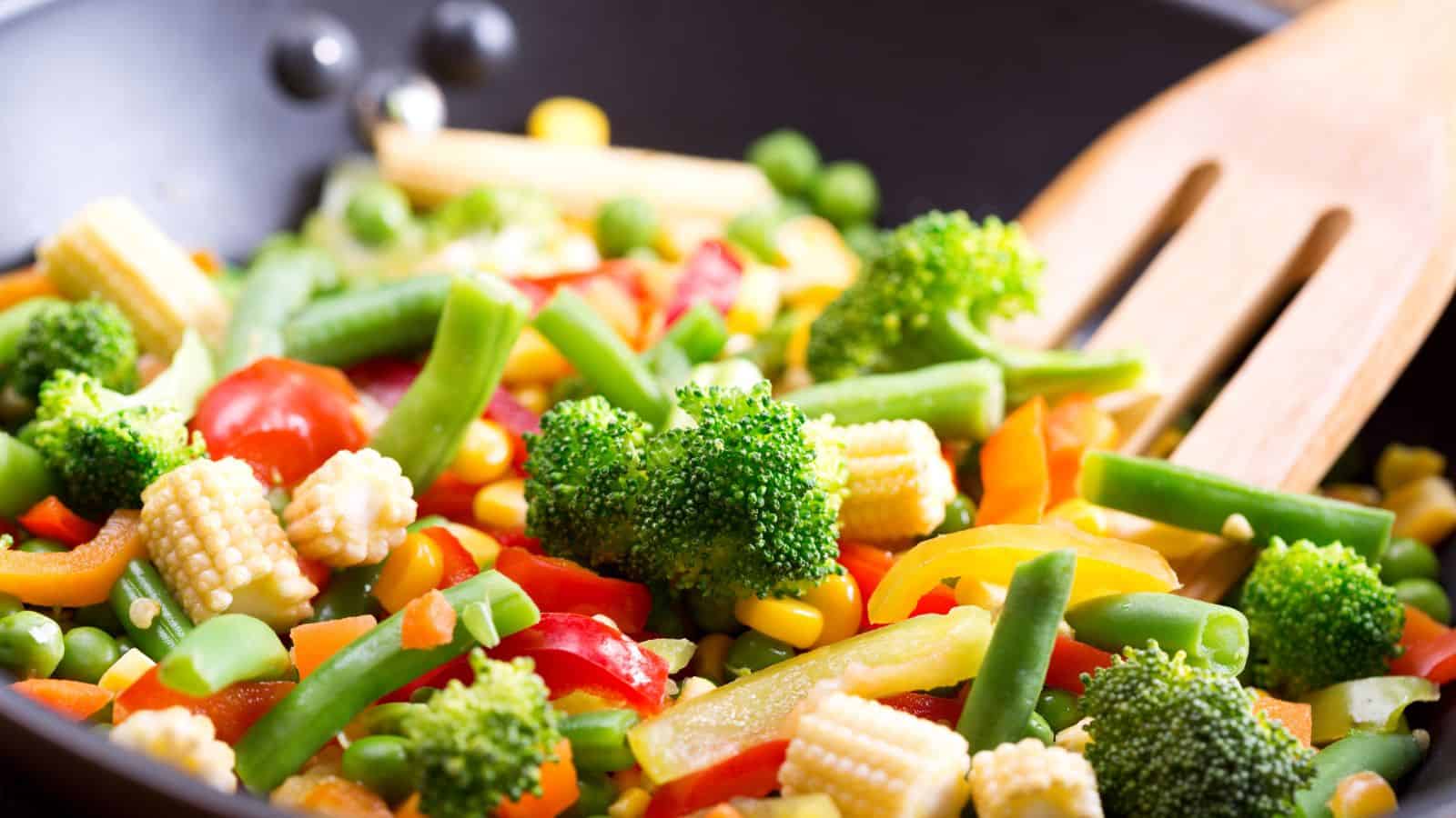 A colorful stir-fry dish with broccoli, baby corn, green beans, orange and red bell peppers, and peas in a black pan. A wooden spatula rests on the edge of the pan, indicating the dish is being prepared or served.