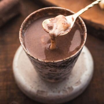A ceramic cup filled with thick, creamy Colombian hot chocolate is placed on a circular ceramic coaster. A spoon, lifting some of the rich chocolate, rests above the cup. The background consists of a wooden surface with blurred elements.