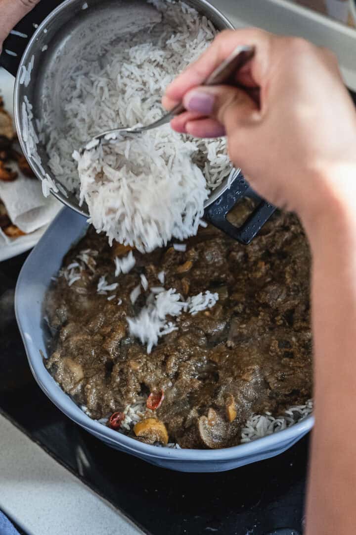 A person is adding cooked white rice from a pot into a bowl on the stove, creating a vibrant vegetarian dish. The mixture includes savory sauce and pieces of zucchini and tomatoes, resembling a delightful biryani-inspired medley.