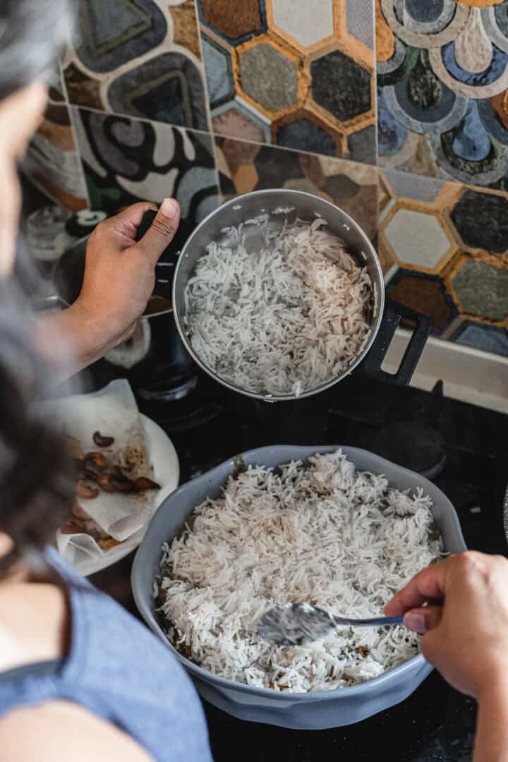 A person spoons cooked white rice from a pot into a light blue baking dish filled with more rice. The vibrant kitchen backdrop features a multicolored hexagonal tile backsplash and a countertop adorned with plates and cooking items, creating the perfect setting for preparing a delightful vegetarian biryani.