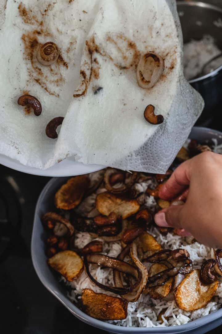 A person gently adds fried onions and mushrooms to a dish of cooked rice in a gray bowl, crafting a simple vegetarian meal. A white paper towel with food remnants hovers above, capturing the essence of homemade comfort.