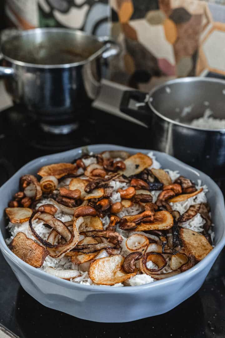 A bowl of fragrant biryani rice, topped with fried onions and sliced mushrooms, sits enticingly on the stovetop. In the background, two pots with handles share the space; one shows its top while the other is partially hidden under a lid. The wall showcases a colorful pattern.
