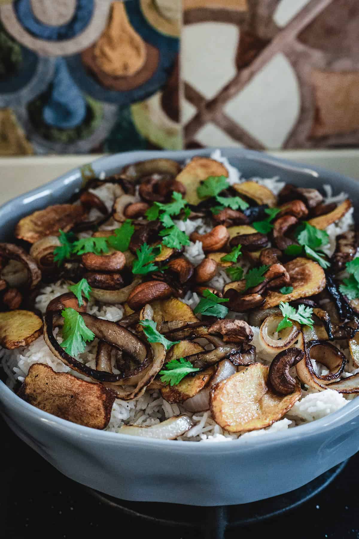 A bowl of vegetarian biryani topped with fried onions, roasted cashews, and potato slices. Fresh cilantro leaves are scattered on top. The dish sits on a dark surface with a colorful tiled background, exuding an inviting aroma.
