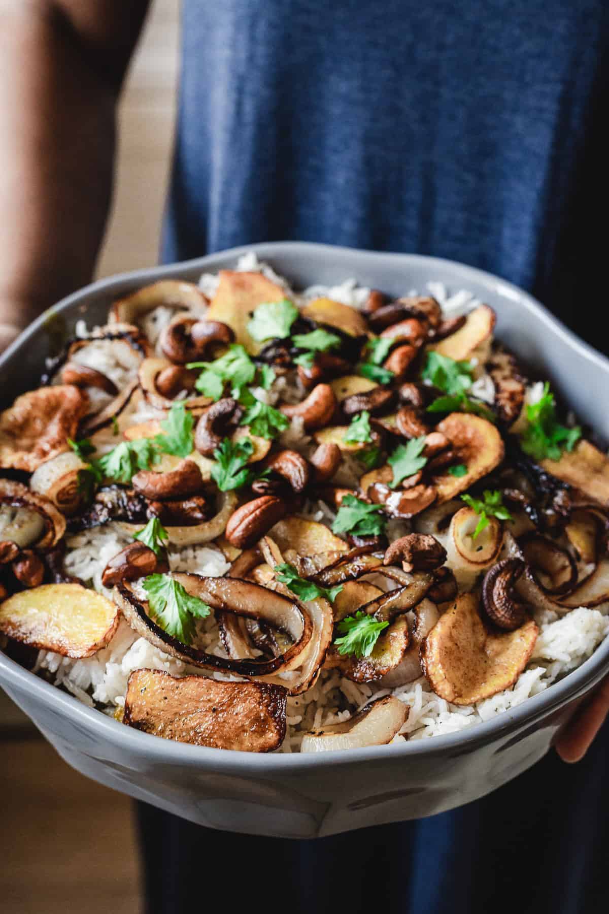 A person holds a bowl of vegetarian biryani, filled with fragrant rice and topped with roasted potatoes, cashews, caramelized onions, and chopped cilantro. The person's upper body is partially visible, dressed in a blue garment.