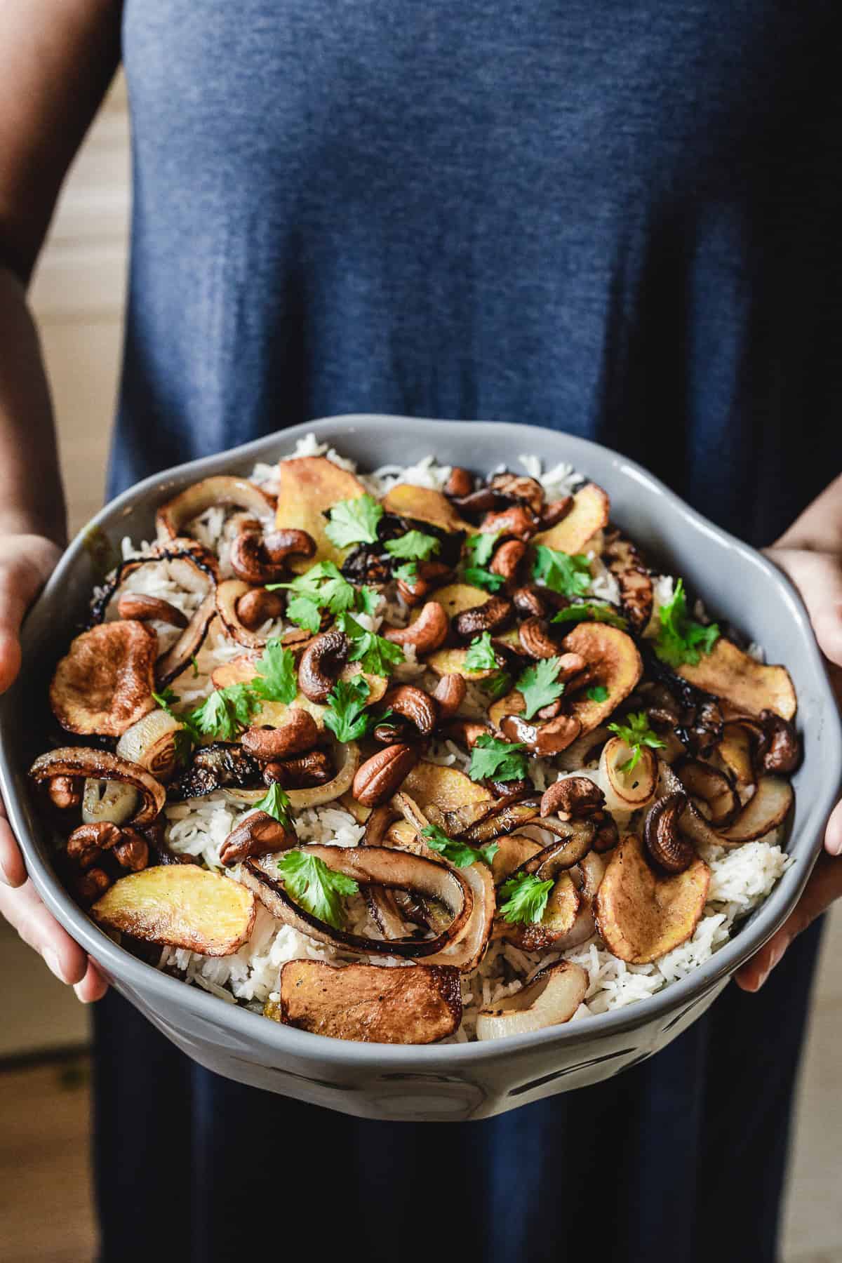 A person in a dark shirt holds a large bowl filled with aromatic biryani, crowned with roasted mushrooms, cashews, and slices of fried onions. Fresh cilantro elegantly garnishes the vegetarian delight.