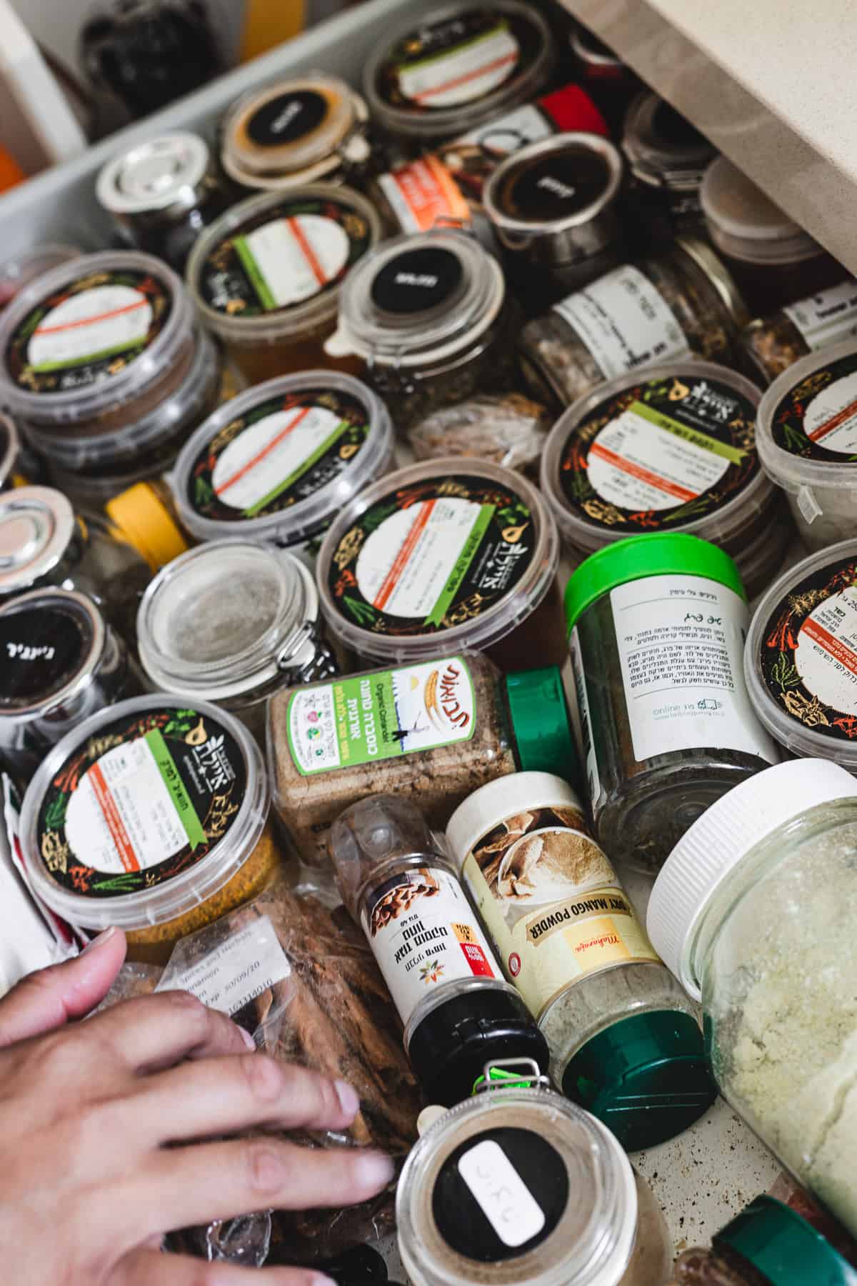 A hand reaches into a drawer filled with various spice jars and containers, each essential for crafting the perfect biryani. Some jars have clear lids, revealing different spices, while others have labels. The neatly arranged containers showcase a vibrant blend of textures and colors.