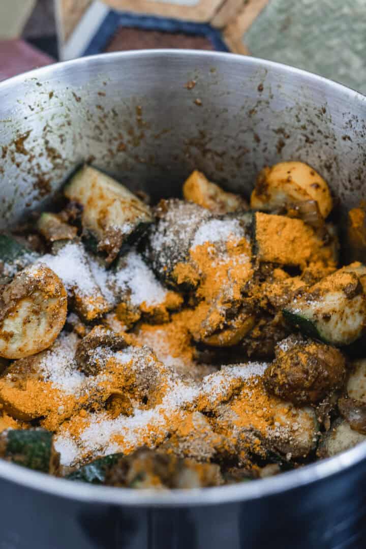 A metal pot contains cut vegetables coated in spices, reminiscent of a vegetarian biryani. Visible ingredients include sliced zucchini and more, with a mix of orange and white powders that appear to be seasoning or spices, missing only the fragrant layer of rice to complete the dish.