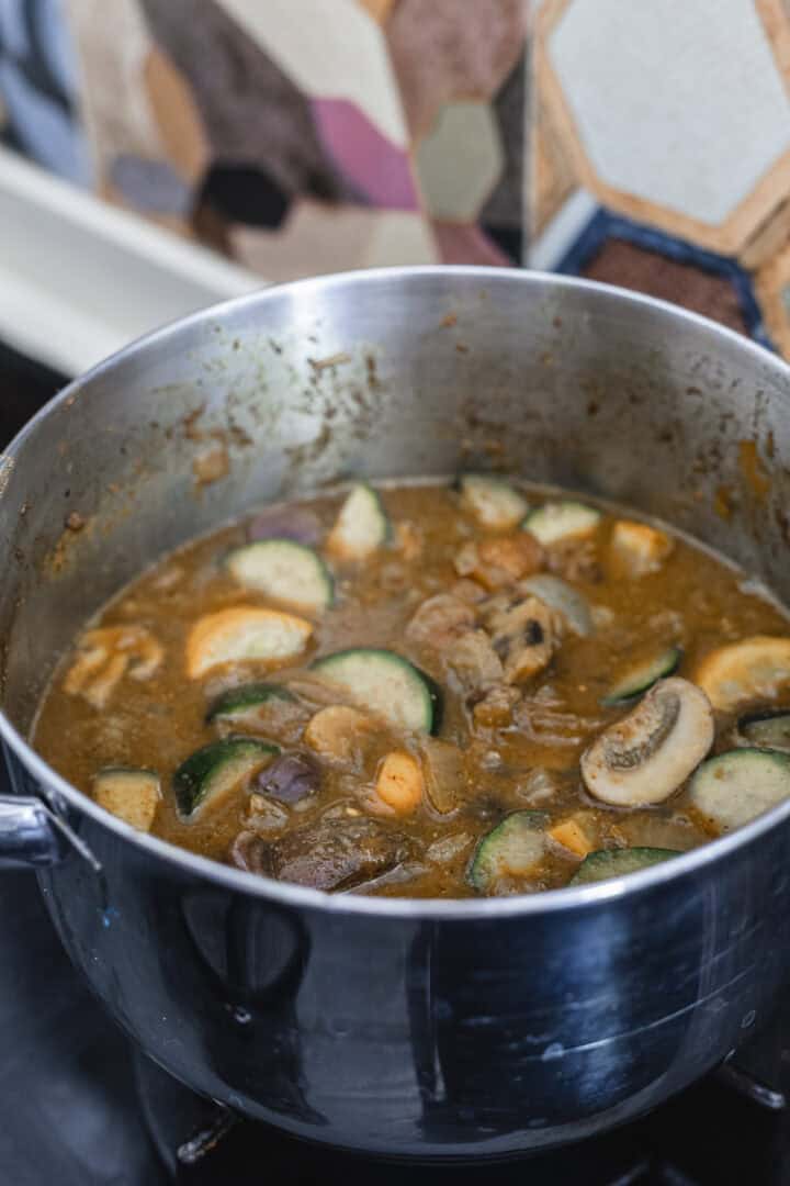 A pot of vegetarian stew containing sliced mushrooms, zucchini, and other vegetables simmers in a rich brown broth on the stove. A geometrically patterned backsplash provides a stylish backdrop.