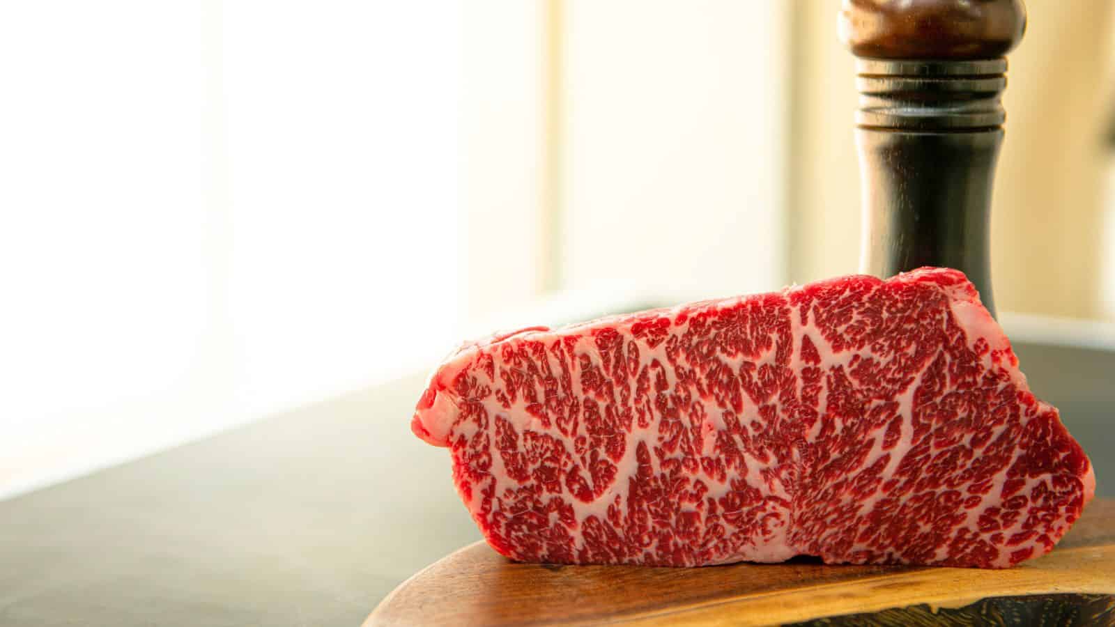 A raw marbled steak rests on a wooden cutting board. A pepper grinder is partially visible in the background. The bright light from the window creates a contrast with the rich red and white marbling of the meat.