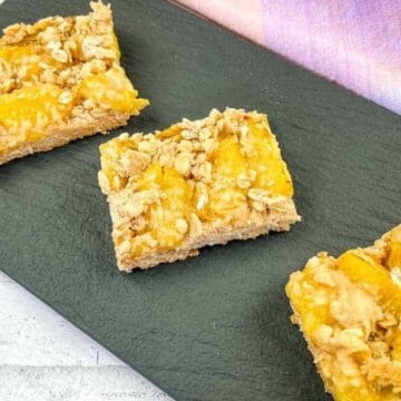 Three square lemon crumb bars are arranged on a dark slate board. The bars have a crumbly topping and a visible lemon layer. The board is placed on a white surface with a pink and purple cloth nearby.