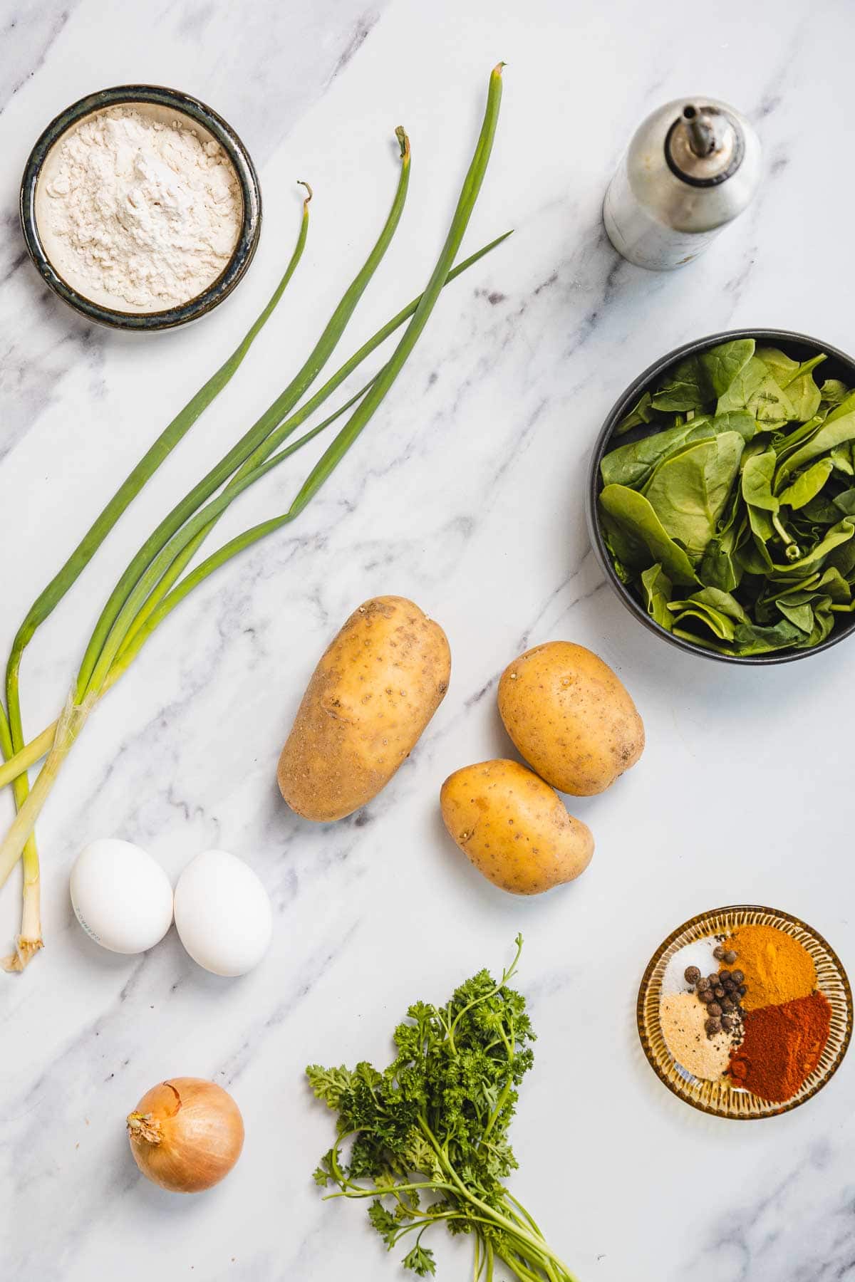 A flat lay of ingredients for Aruk, Iraqi style latkes, on a marble surface&mdash;green onions, spinach, potatoes, flour, eggs, brown onion, parsley, assorted spices, and a metal shaker.