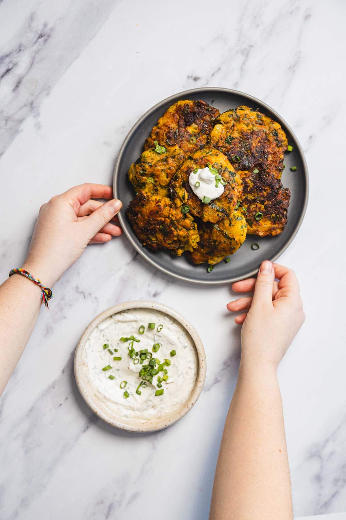 A person holds a gray plate with several golden-brown Aruk, or Iraqi Style Latkes, topped with a dollop of white sauce and chopped green onions; a bowl of dip garnished with green onions sits nearby on a marble surface.