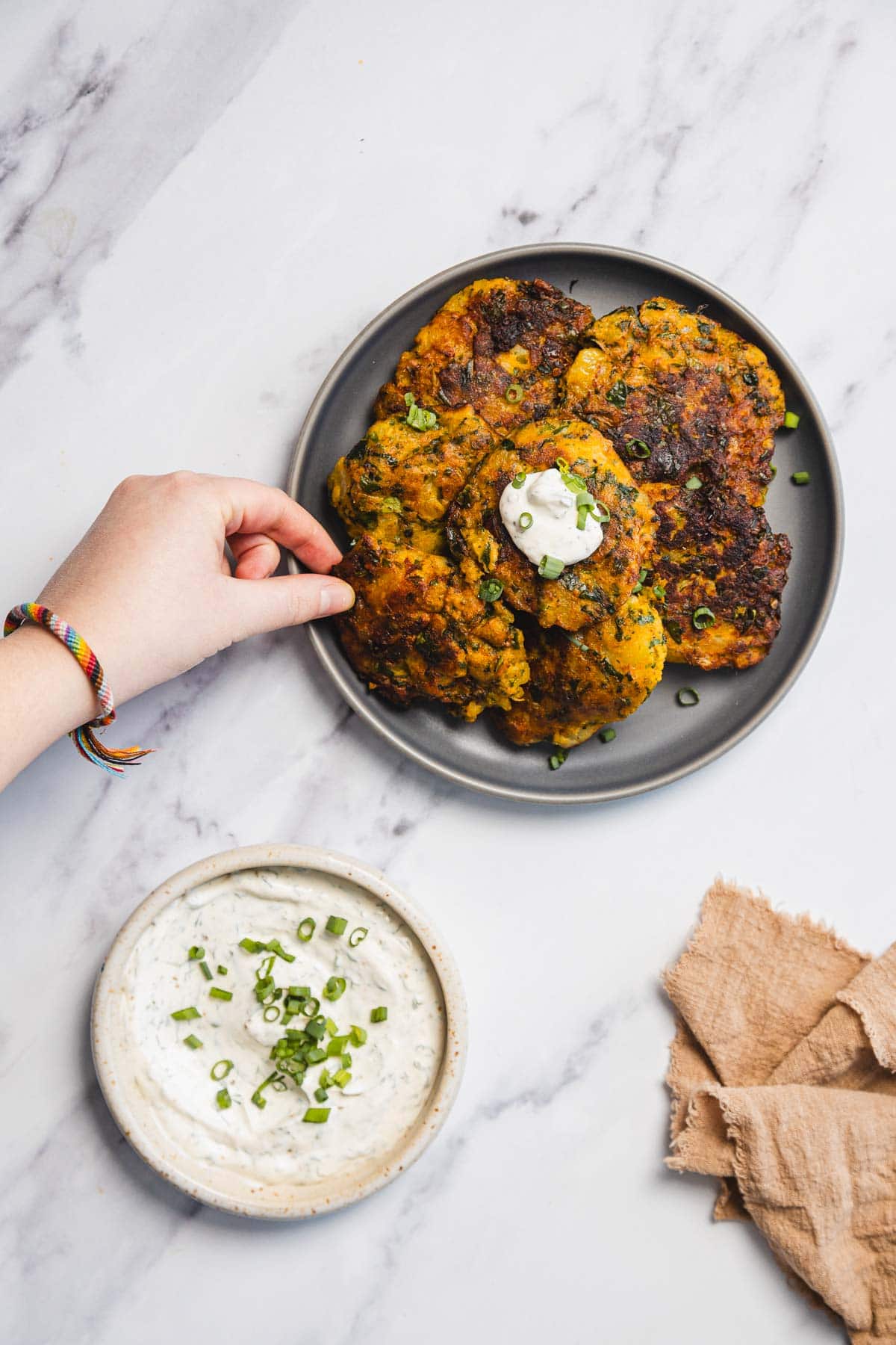 A hand reaches for an Iraqi style vegetable fritter, or Aruk, on a gray plate topped with sour cream and chopped green onions; a bowl of dipping sauce and a beige napkin rest nearby on a marble surface.
