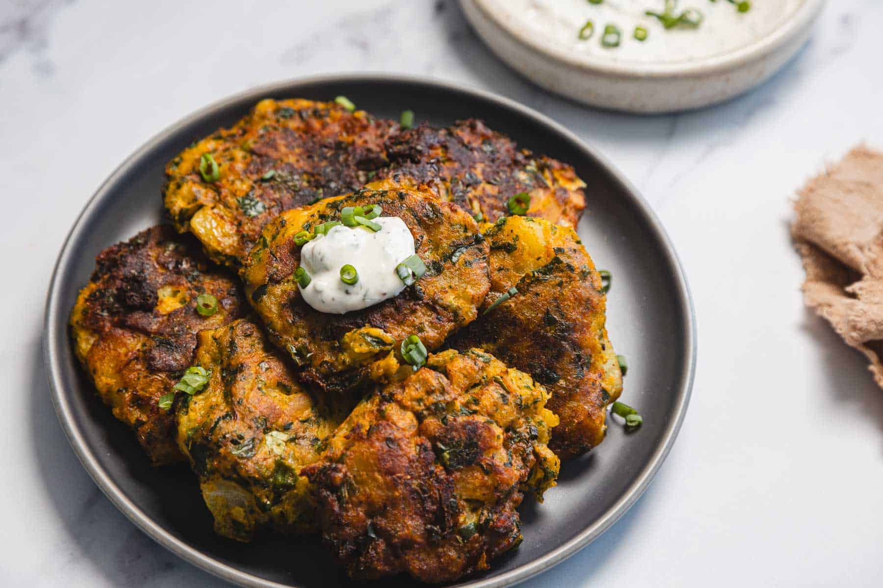 A dark plate holds a stack of golden-brown Aruk, Iraqi Style Latkes, garnished with chopped green herbs and topped with a dollop of white sauce. A bowl of similar sauce is visible in the background.