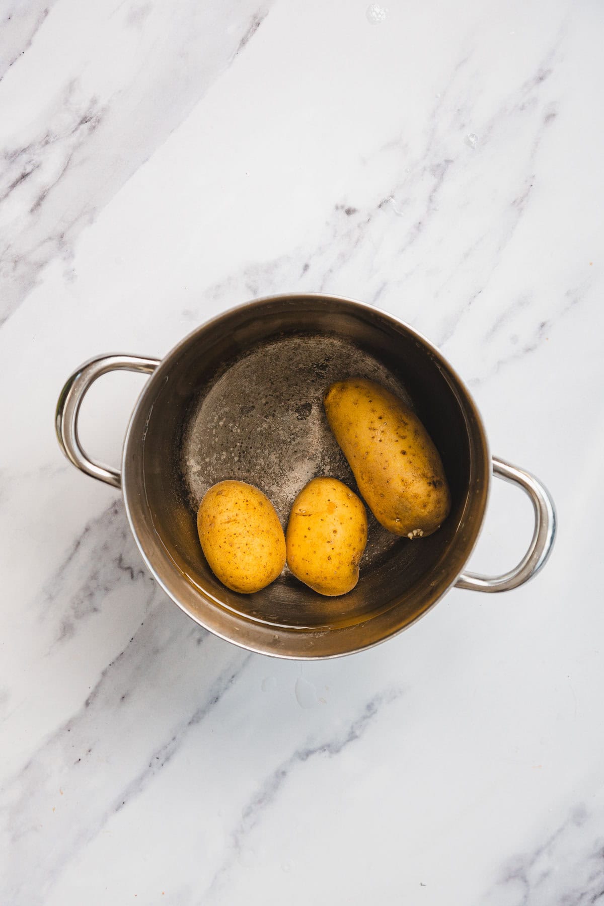 A stainless steel pot containing three unpeeled potatoes sits on a white marble surface. The pot, with some water at the bottom, holds the potatoes often used in Aruk, a beloved dish in Iraqi cuisine similar to latkes.