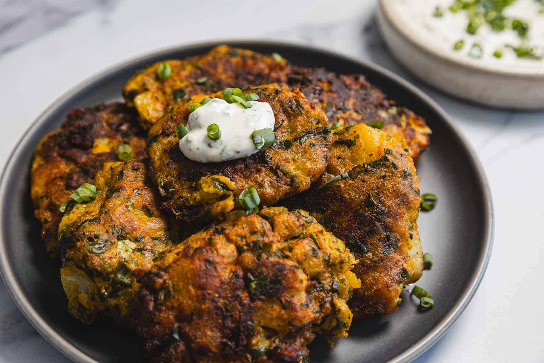 A black plate holds several golden-brown Aruk, or Iraqi Style Latkes, garnished with chopped green onions and a dollop of white sauce. A small bowl of sauce with green herbs is visible in the background.
