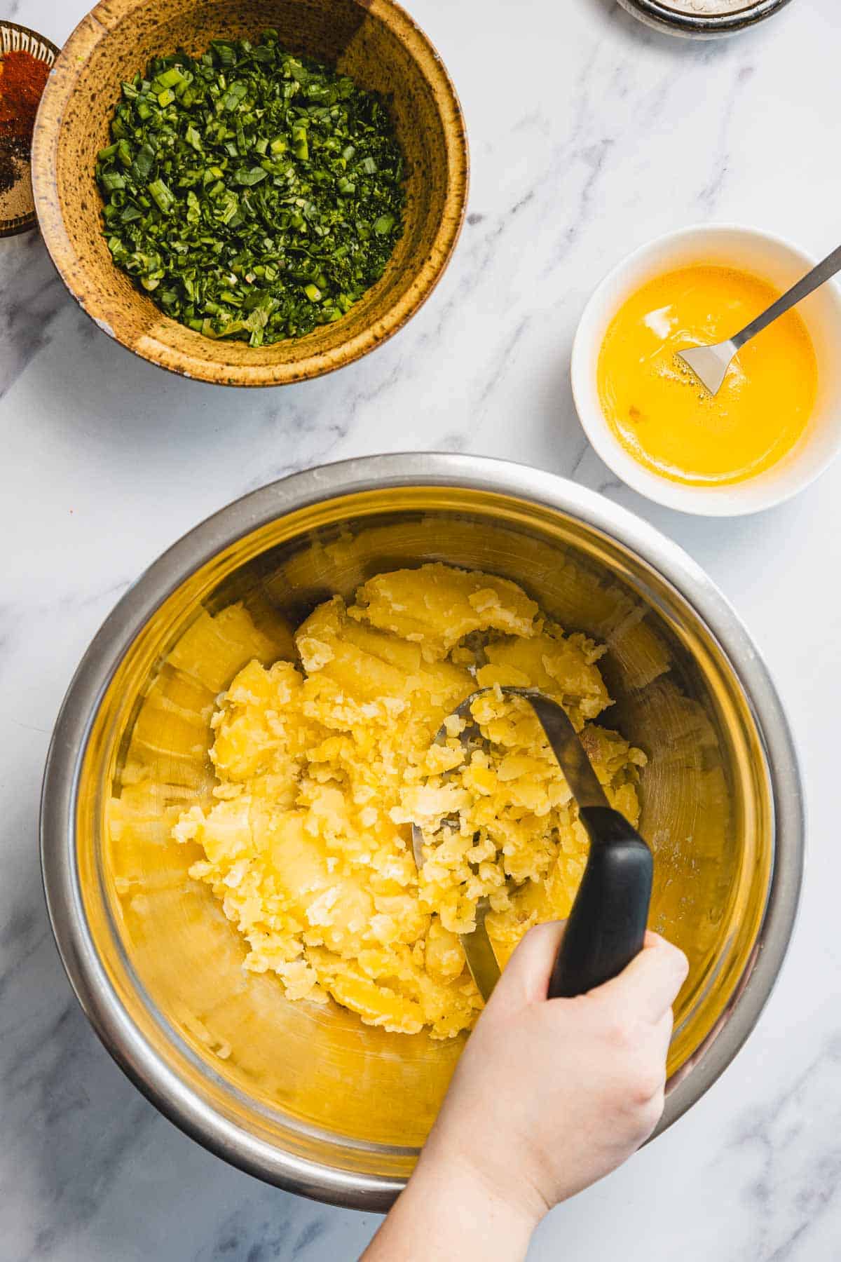 A hand mashing boiled potatoes in a metal bowl, preparing Aruk, Iraqi style latkes, with bowls of chopped greens and beaten egg nearby on a marble countertop.
