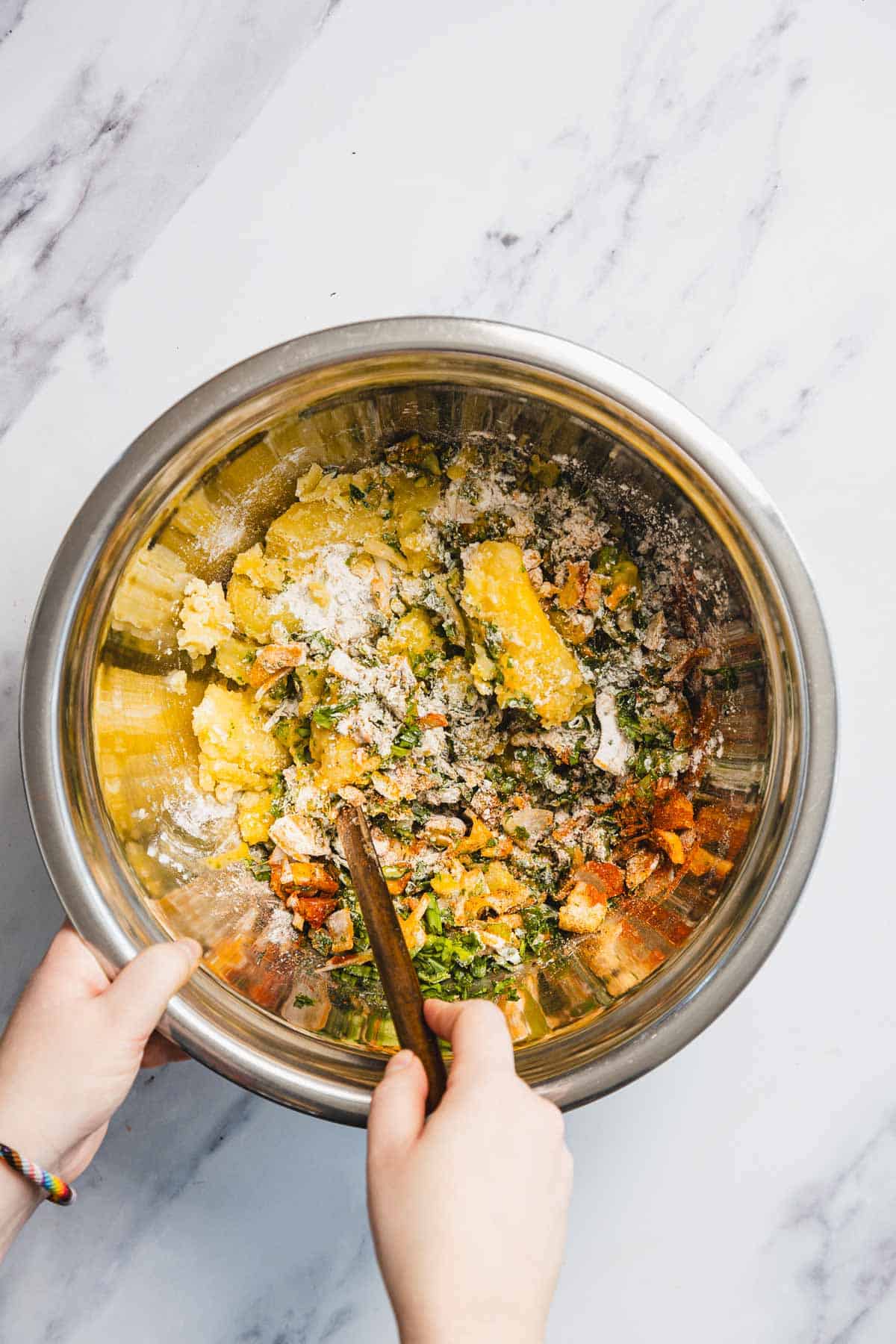 A person uses a wooden spoon to mix vegetables, flour, and other ingredients for Iraqi style latkes, also known as Aruk, in a large stainless steel bowl on a marble surface.