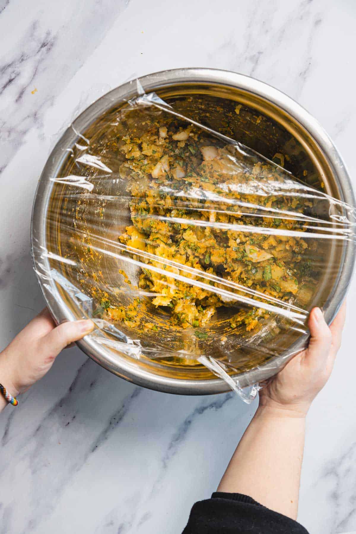 A person covers a metal mixing bowl containing a yellow and green vegetable mixture for Aruk, delicious Iraqi latkes from Middle Eastern cuisine, with plastic wrap on a white marble countertop.
