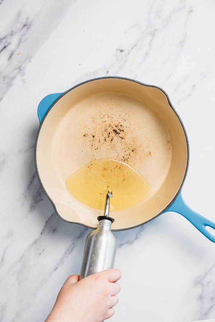 A hand holds a metal oil dispenser, pouring oil into a large, round, beige enameled cast iron skillet with a blue handle on a white marble surface, ready for frying Iraqi style latkes known as Aruk.