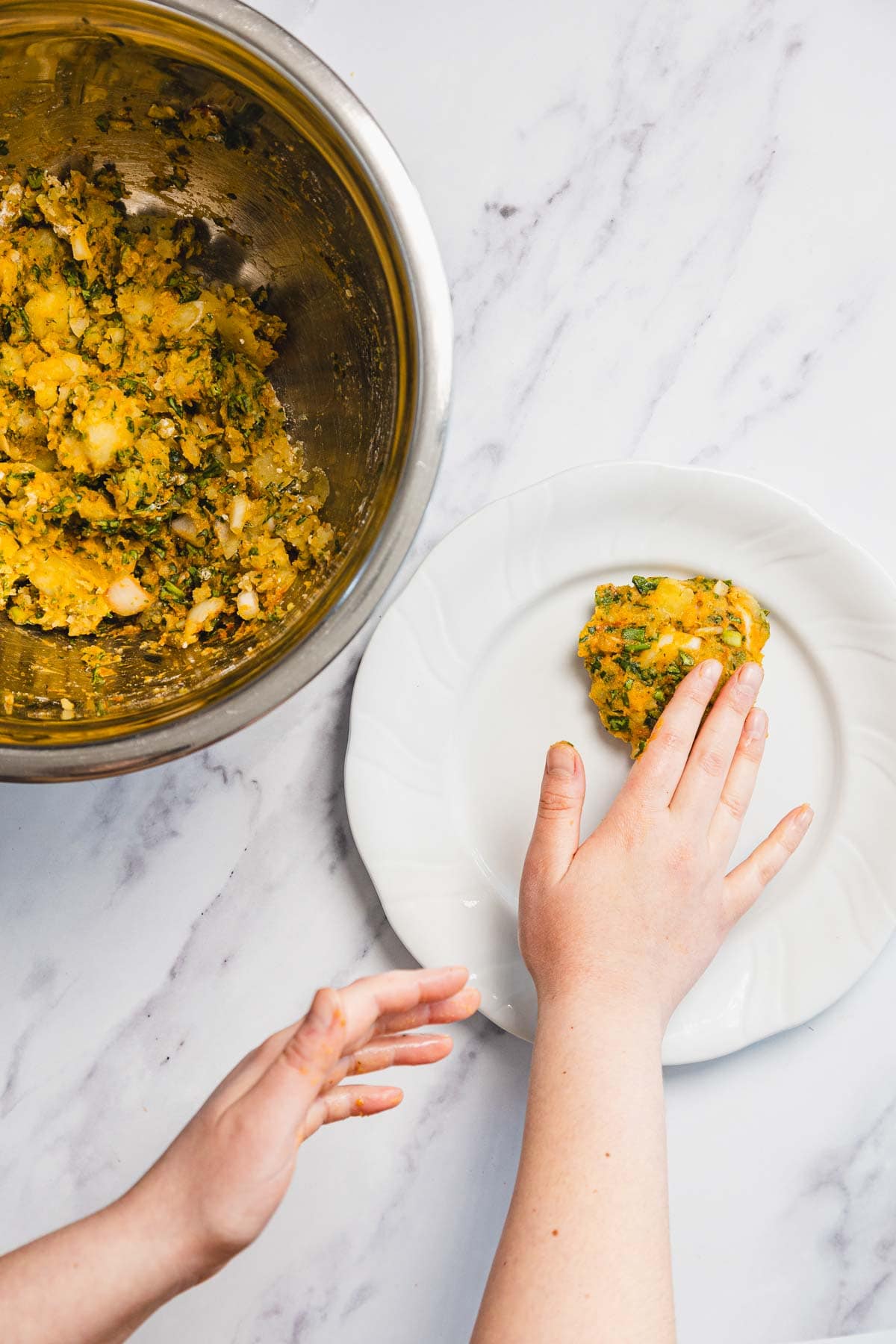 A person presses a patty-shaped portion of Aruk, an Iraqi Style chickpea and herb mixture, onto a white plate. A metal bowl with more of the mixture sits nearby on a marble countertop.