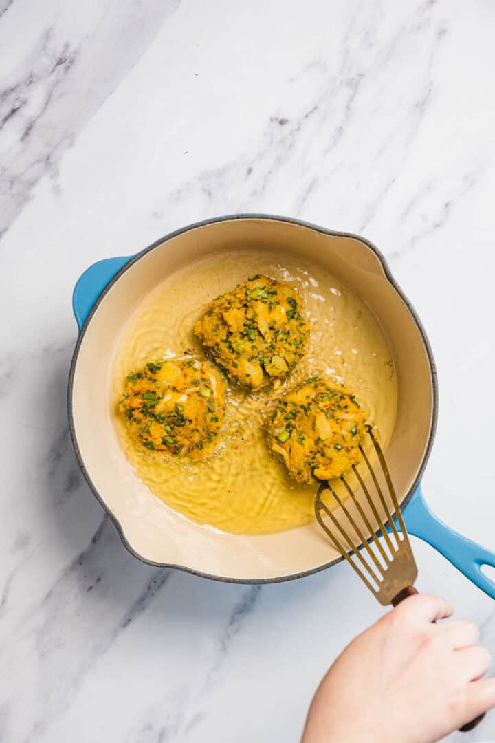 A hand uses a slotted spatula to fry three round Iraqi style latkes in a light blue pot filled with oil, on a marble countertop.