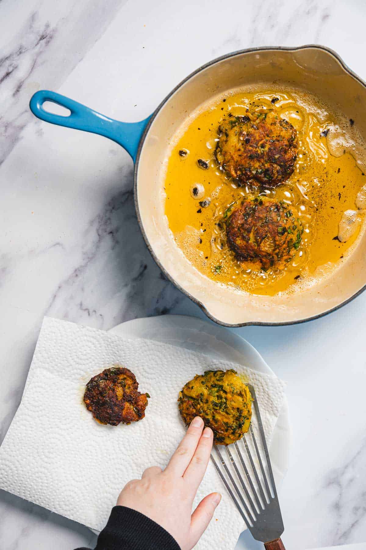 A hand places an Iraqi style latke, known as Aruk, onto a paper towel-lined plate next to a skillet with two more patties frying in oil. The scene is set on a marble countertop.