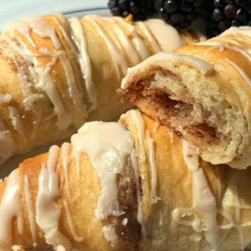 Three crescent rolls drizzled with white icing are arranged on a white plate with a few blackberries in the background. One of the rolls is cut open, showing a cinnamon filling.