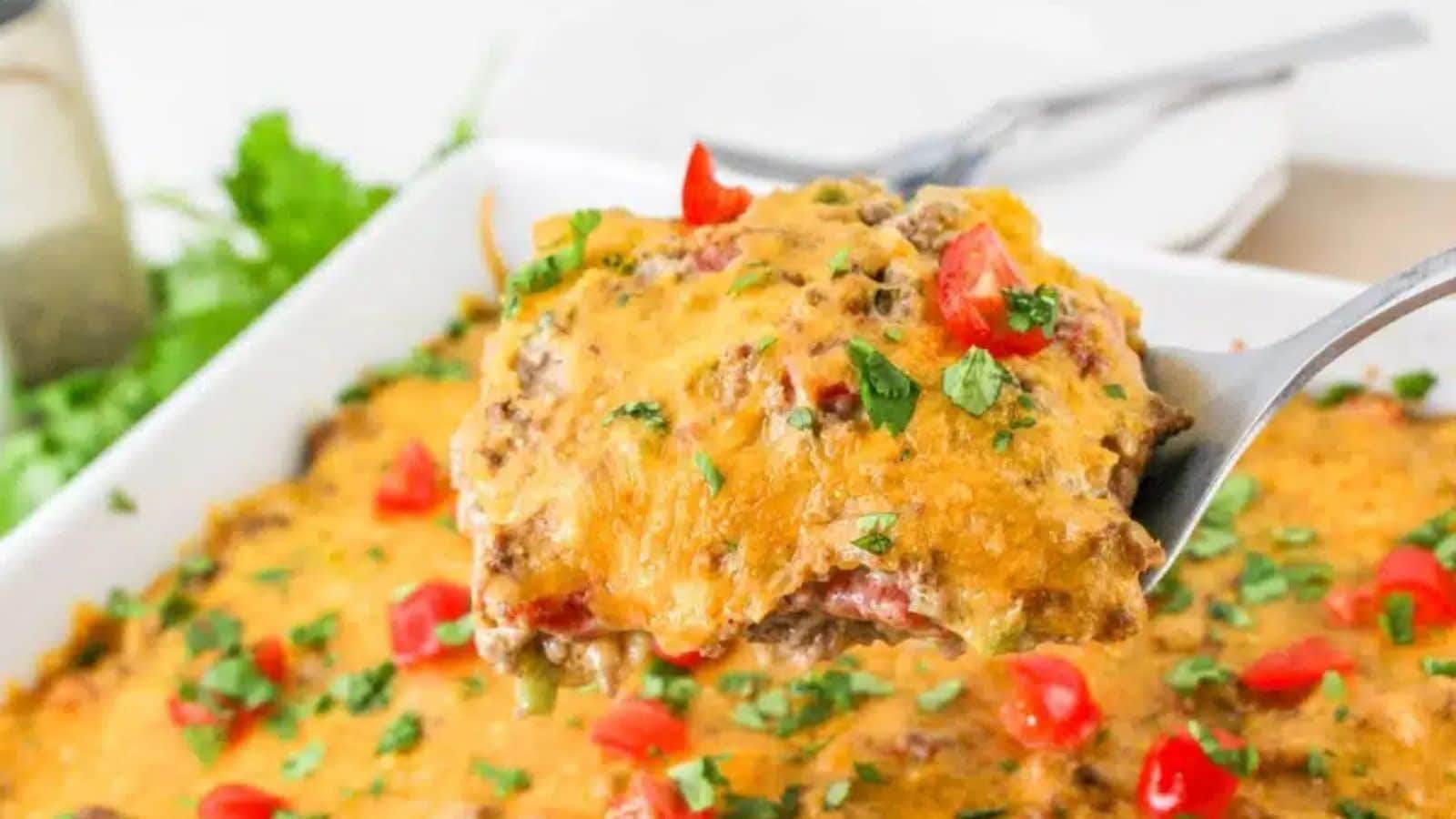 Close-up of a spoon lifting a portion of cheesy beef casserole garnished with chopped tomatoes and herbs from a white baking dish.