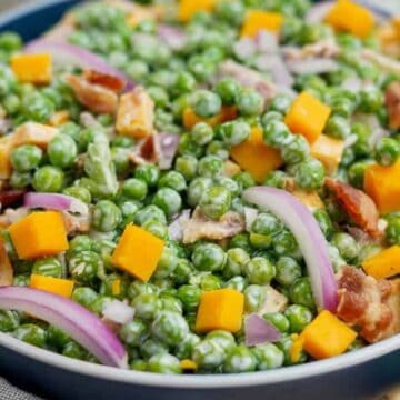 A bowl filled with a salad made of green peas, diced cheddar cheese, red onion slices, bacon pieces, and a creamy dressing, set on a woven placemat.