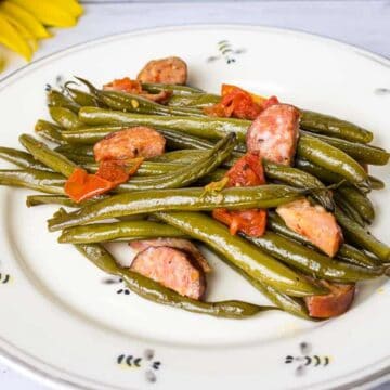 A white plate with cooked green beans, sliced sausage, and diced tomatoes. The plate is on a wooden surface next to a sunflower.