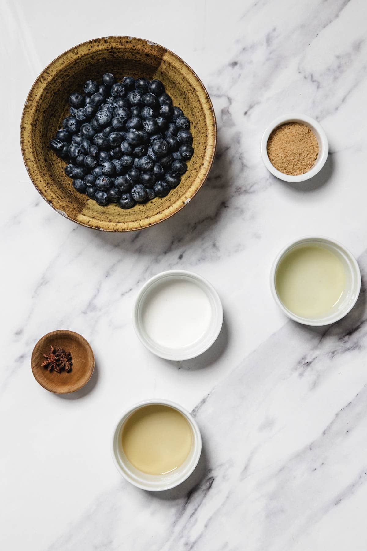 A bowl of blueberries, small bowls containing brown sugar, white liquid, yellow liquid, clear liquid, and a small wooden bowl with whole cloves are arranged on a white marble surface—perfect ingredients for making homemade blueberry sauce.
