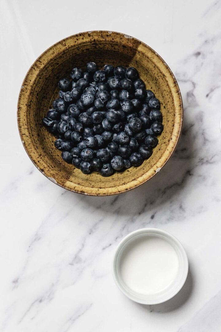 A brown ceramic bowl filled with fresh blueberries sits on a white marble surface next to a small white cup of milk or cream, perfect for making homemade blueberry sauce.
