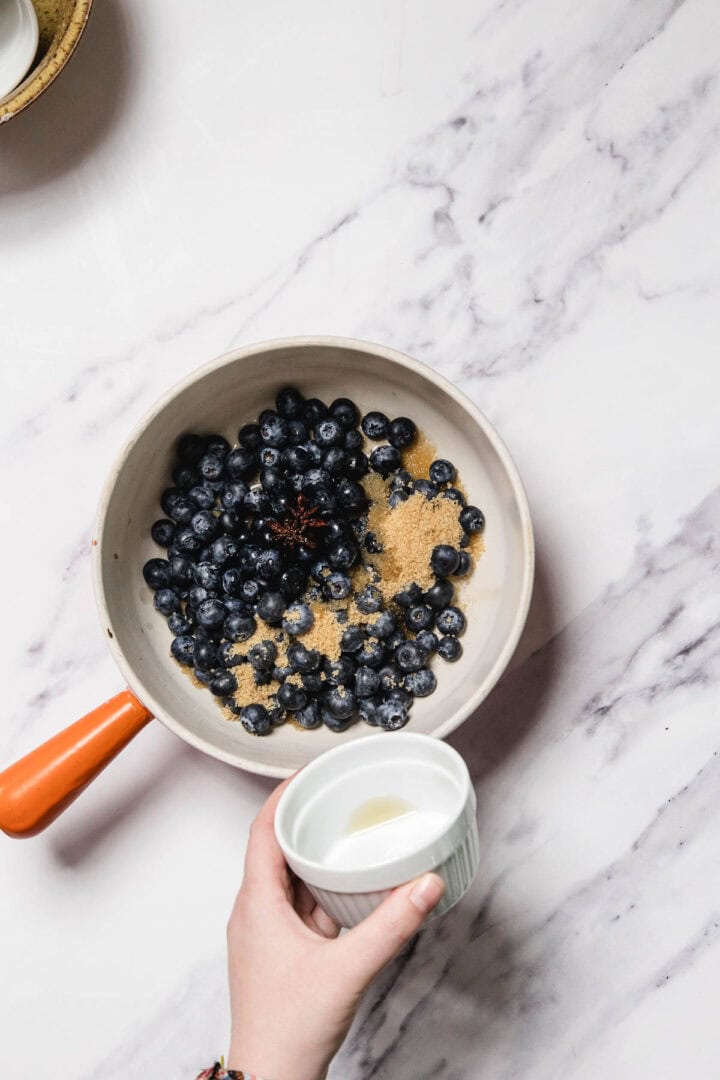 A hand holds a small white ramekin filled with liquid above a saucepan of blueberries and brown sugar, preparing to craft homemade Blueberry Sauce on a marble countertop.