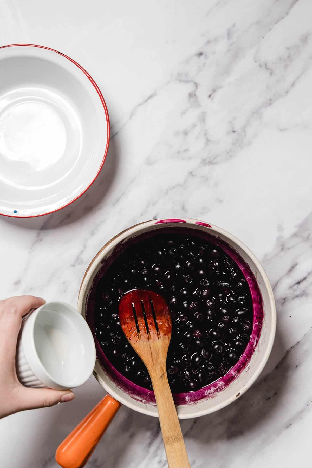A hand holds a small white bowl above a saucepan filled with blueberry sauce made from cooked blueberries, sitting on a marble countertop. A wooden spoon rests in the saucepan, and an empty white bowl is nearby.